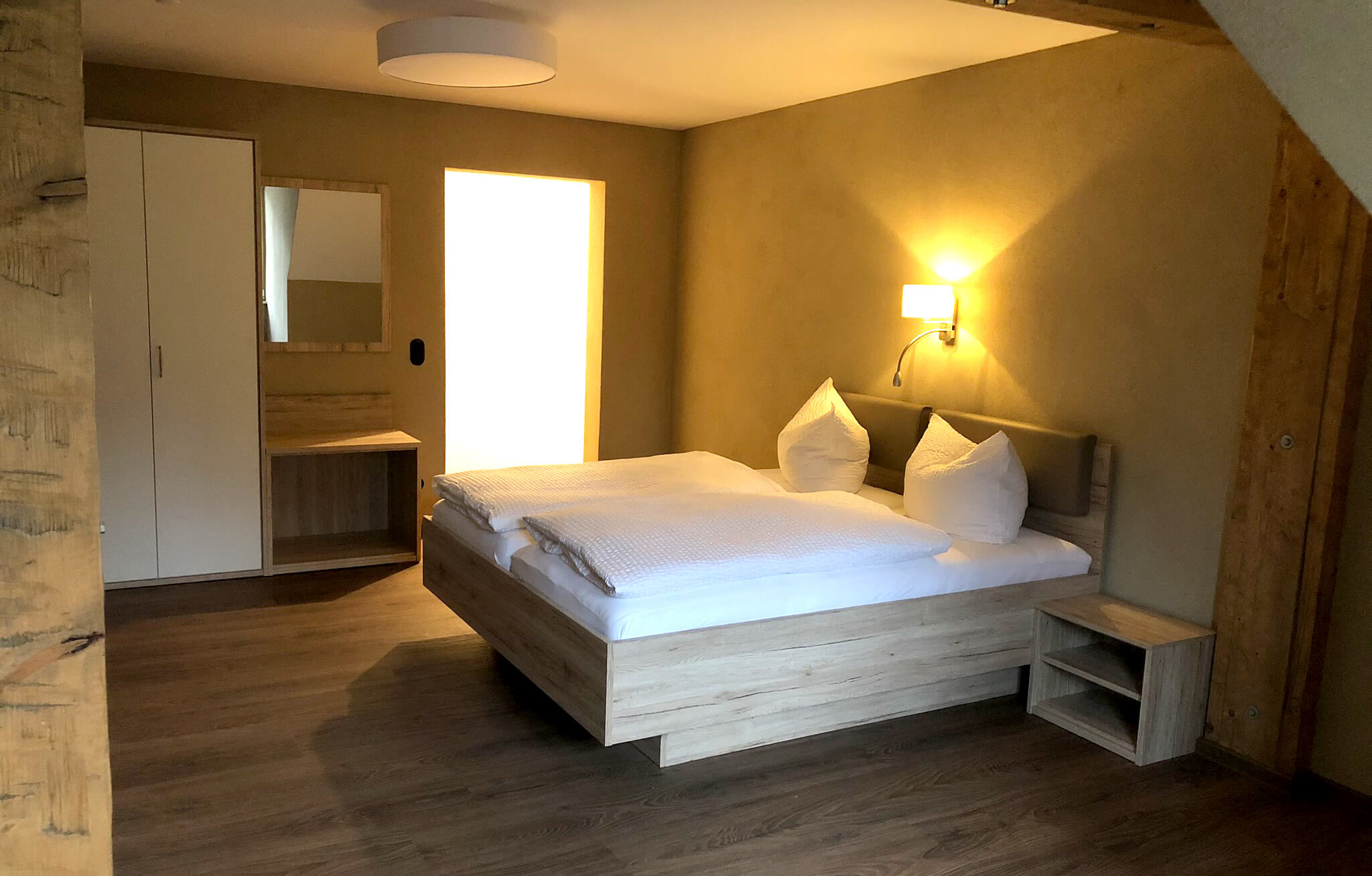 Hotelzimmer mit großem Doppelbett und Einbauschrank. Über dem Bett hängt eine Wandleuchte und hüllt das bett in ein warmes Licht.
